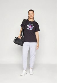 Mujer con camiseta gráfica negra, pantalones deportivos blancos, zapatillas blancas, llevando un bolso negro al hombro, de pie contra un fondo claro liso.