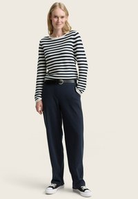 Femme debout portant un haut à rayures noir et blanc à manches longues, une ceinture noire, un pantalon large bleu marine et des baskets noir et blanc sur un fond neutre.