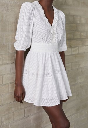 Kobieta w białej teksturowanej mini sukience z bufkami i falbaniastym dekoltem w kształcie litery V, stojąca na tle beżowej ceglanej ściany.