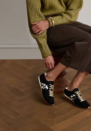 Black suede sneakers with white and black spotted accents, featuring beige laces and a light-colored sole, set against a wood floor.