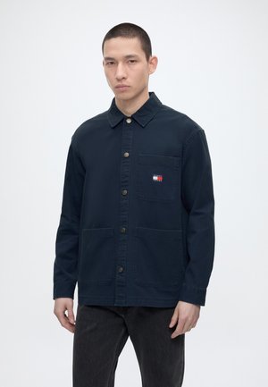 Νέος άνδρας που φοράει σκούρο μπλε μπουφάν με κουμπιά και τσέπες στο στήθος και κάτω, στέκεται μπροστά σε απλό φωτεινό φόντο.