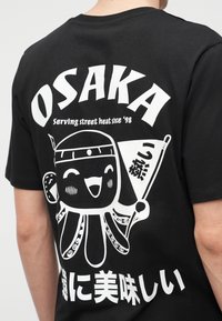 Μαύρο T-shirt με λευκό καρτουνίστικο χταπόδι που κρατάει σημαία, το κείμενο "OSAKA" από πάνω και ιαπωνικούς χαρακτήρες από κάτω, φορεμένο από ένα άτομο που φαίνεται από πίσω.