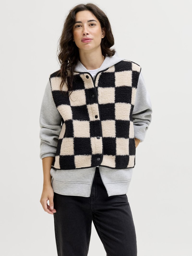 Femme aux cheveux foncés portant une veste polaire à carreaux noire et beige par-dessus un sweat à capuche gris clair zippé et un pantalon noir, debout devant un fond uni.