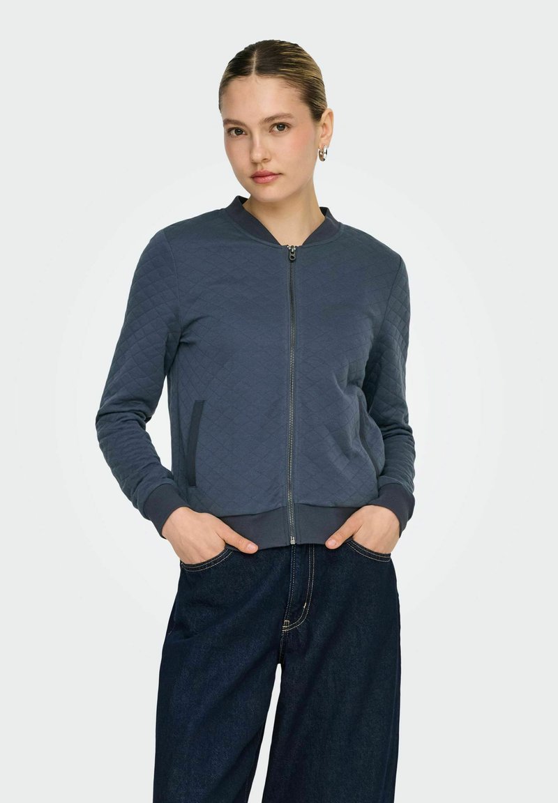 Femme aux cheveux lissés en arrière portant une veste matelassée bleu marine zippée et un jean large bleu foncé, debout les mains dans les poches.
