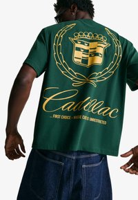 Green short-sleeve t-shirt featuring a large gold Cadillac logo and text on the back, with a relaxed fit and smooth cotton texture.