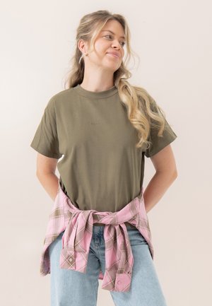 Kvinde med langt blondt hår iført en olivengrøn t-shirt, lyseblå jeans og en lyserød ternet skjorte bundet rundt om taljen, kigger til siden.