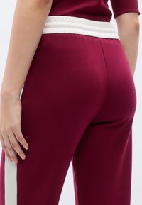 Μπορντό αθλητικό παντελόνι με λευκή ελαστική ζώνη μέσης, με λεία υφή και λευκή ρίγα στο πλάι. Κοντινή όψη από πίσω.