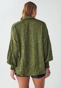 Oliwkowa, oversize'owa kurtka o pomarszczonej fakturze, z szerokimi rękawami i ściąganymi mankietami. Posiada prosty kołnierz i luźny krój.