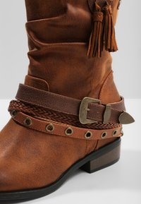 Stivale in pelle marrone con design morbido, dettagli in cinturino intrecciato, decorazioni in metallo rotondo e un piccolo tacco impilato.