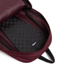 L'intérieur du sac à dos marron présente un motif à carreaux noirs, un matériau synthétique, une fermeture éclair et une étiquette logo à la base.