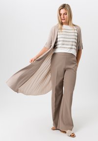 Cardigan long beige avec dos plissé, chemise rayée blanche et beige, et pantalon large beige. Sandales marron à enfiler avec un design tissé.