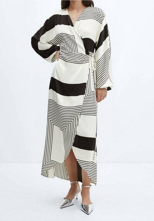 Femme portant une robe portefeuille à motif géométrique noir et blanc avec des manches longues bouffantes et des talons pointus argentés sur un fond uni.