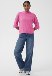 Rosa Pullover mit hohem Kragen und kurzen Ärmeln, kombiniert mit weit geschnittenen blauen Jeans. Das Outfit zeichnet sich durch eine entspannte Passform und eine weiche Stoffstruktur aus.