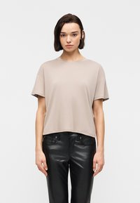 Γυναίκα με κοντά σκούρα μαλλιά που φορά χαλαρό μπεζ κοντομάνικο τοπ και μαύρο δερμάτινο παντελόνι, στέκεται μπροστά σε απλό λευκό φόντο.