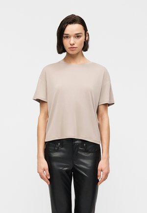 Femme aux cheveux courts et foncés portant un haut ample beige à manches courtes et un pantalon en cuir noir, debout devant un fond blanc uni.