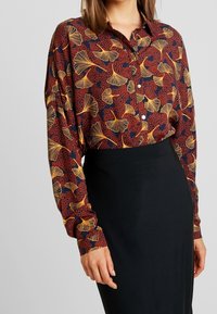 Chemise à manches longues avec boutons, en tissu rouge foncé orné d'un motif de feuilles de ginkgo jaunes, associée à une jupe noire cintrée. Texture douce.