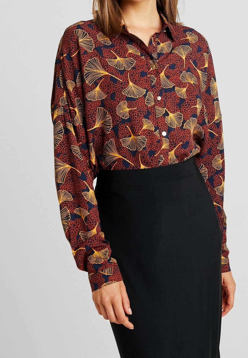 Chemise à manches longues avec boutons, en tissu rouge foncé orné d'un motif de feuilles de ginkgo jaunes, associée à une jupe noire cintrée. Texture douce.