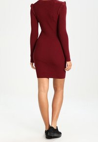 Abito a coste color bordeaux con maniche lunghe, silhouette aderente, dettagli arricciati sulle spalle, lunghezza al ginocchio, indossato con scarpe con platform nere.