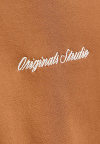 Tessuto marrone con finitura testurizzata che presenta un testo ricamato bianco con la scritta "Originals Studio." Dettaglio visivo chiave è la morbidezza della texture.