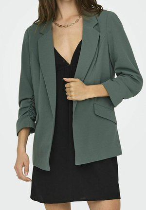 Mujer con blazer verde oscuro con mangas fruncidas sobre un vestido negro con escote en V y un collar de cadena dorada, mano ajustando el blazer.
