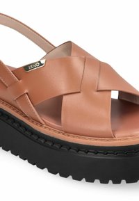 Sandale en cuir brun clair avec design croisé, bout ouvert, et une semelle en caoutchouc noire épaisse présentant un motif texturé. Accent de logo doré visible.