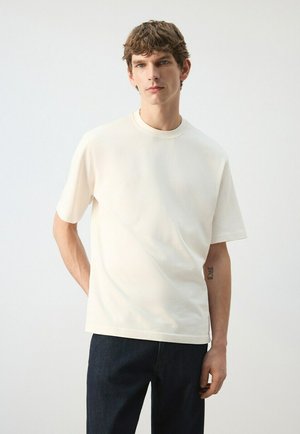 Jonge man met kort bruin haar draagt een effen wit T-shirt met korte mouwen en donkere spijkerbroek, staat met één hand achter zijn rug.