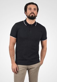 Černé polokošile s krátkým rukávem z texturované látky, s klasickým límcem a bílými akcenty podél límce a rukávů.
