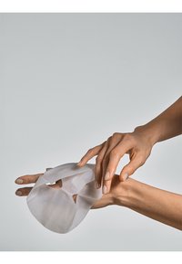 Clear facial mask held by a hand, made of translucent, textured material with eye and mouth openings. Natural nail polish on fingers.