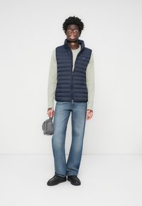 Marine puffervest over een lichtgroen langemouwen shirt, gecombineerd met blauwe flared jeans en zwarte schoenen; houdend een kleine grijze tas.