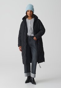 Schwarzer, gesteppter langer Mantel mit Reißverschluss, grauer Knopf-Sweater, dunkle Jeans mit umgeschlagenen Säumen und eine hellblaue Strickmütze.