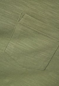 Tejido verde con una textura sutil presenta un bolsillo de parche cosido con hilo a juego. El diseño es simple y funcional.