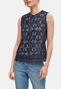 Marineblaues, ärmelloses Oberteil aus Spitzenstoff mit floralen Mustern, gewelltem Saum und rundem Halsausschnitt mit kontrastierendem Besatz. Kombiniert mit hellblauen Jeans.
