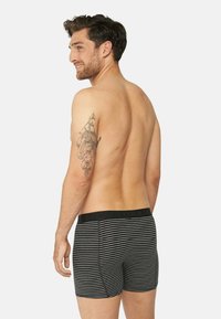 Pantaloni boxer a righe bianco e nero con vita nera solida e lettere in rilievo. Il modello ha un tatuaggio sul braccio.