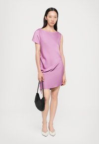 Robe courte violette à coupe décontractée, décolleté bateau et manches courtes. Le modèle tient un sac à main tissé noir et porte des chaussures pointues beiges.
