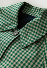 Tessuto a quadri verde e beige con scollo a collo, caratterizzato da un bottone scuro e una trama liscia. Vista ravvicinata dei dettagli del design.