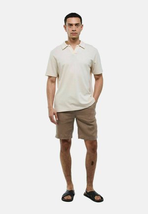 Polo shirt crème avec un col en V, associé à un short taupe et des sandales noires. Le tissu semble lisse et la coupe est décontractée.