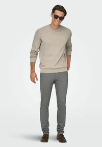 Helebeež tekstuuriga kudumchi bleiser, mida on kombineeritud hallide kitsaste pükstega. Modell kannab tumedaid päikeseprille, pruune loafer-kingi ja naeratab.