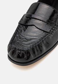 Mocassin en cuir noir brillant avec couture moc toe et un détail de bride horizontale sur la partie supérieure de la chaussure.