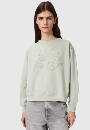 Femme aux longs cheveux bruns portant un sweat-shirt vert clair avec des lettres "AS" texturées et un jean gris délavé, debout devant un fond uni.