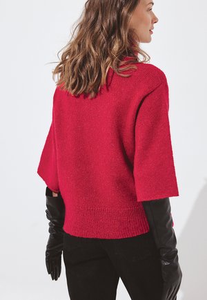 Červený pletený sveter s krátkymi rukávmi, rebrovaným lemom a voľným strihom. Kombinovaný s čiernymi koženými rukavicami. Hladká textúra so subtílnym leskom.