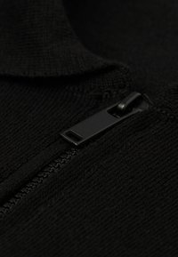 Černá pletená tkanina s texturovaným povrchem; obsahuje černý zip s obdélníkovým poutkem, umístěným na pravé straně oděvu.
