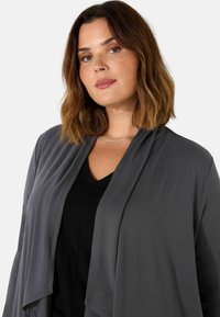Šedý cardigan s otevřeným předním dílem a splývavým designem, vyrobený z lehkého materiálu, který se příjemně nosí přes černý top s výstřihem do V, s hladkou texturou a jemným splýváním.