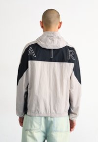 Lightweight jacket in gray and black, featuring an adjustable hood, elastic cuffs, and "AIR" lettering on the back. Smooth, textured fabric.