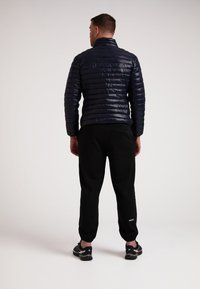 Marineblå polstret jakke med høj krave, lodrette syninger og en blank overflade, parret med sorte joggers og sporty fodtøj.