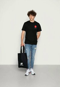 Joven con camiseta gráfica negra, jeans azules rasgados, zapatillas New Balance blancas, sosteniendo una bolsa de mano negra de New Balance, de pie frente a una pared lisa.