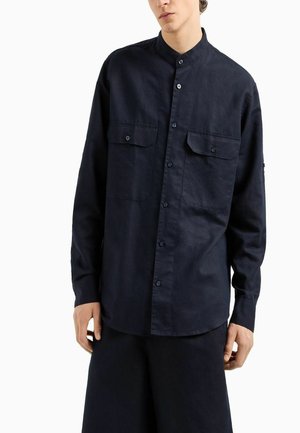 Camicia blu navy a maniche lunghe realizzata in un tessuto con una leggera trama, caratterizzata da una chiusura con bottoni, due tasche sul petto e un colletto alla coreana.