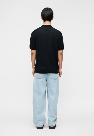 Άτομο που φαίνεται από πίσω, φοράει μαύρο κοντομάνικο πουκάμισο, ανοιχτό μπλε τζιν με φαρδιά πόδια και μαύρα παπούτσια, πάνω σε απλό φόντο.