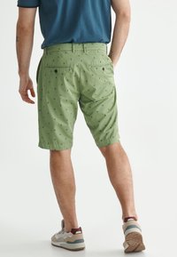 Homme portant un short à motifs verts, une chemise bleue et des baskets beiges, se tenant les mains dans les poches, de dos sur fond blanc.