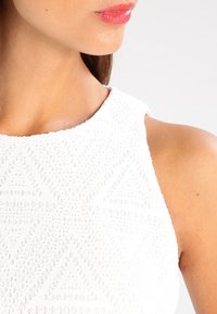 Top sin mangas blanco con un patrón texturizado que presenta diseños geométricos en relieve, un escote alto y una tela suave.