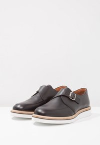 Scarpe in pelle nera con cinturino a fibbia, punta tondeggiante e suola in gomma bianca. Texture liscia con dettagli di cucitura lungo i bordi.